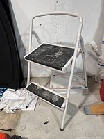 Side angle view of white metal frame two-step folding stepladder with black steps showing paint stains and wear.
