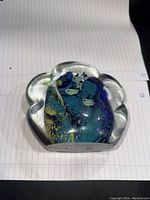 Front view of thick clear glass paperweight with colorful, abstract swirls and shapes inside in blue, yellow, green and white, sitting on white lined paper.