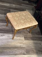 Photo of entire vintage stool showing padded damask fabric seat with richly ornamented gold-painted carved wooden frame and cabriole legs on wooden floor.