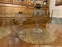 Two oil lamps side by side with various glass chimneys and shades arranged on table