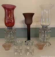 Two hurricane lamps and five small candleholders arranged side by side, showing cranberry and clear glass chimney shades with hanging crystals plus small candleholders in front.