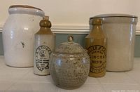 Group photo showing 2 medium stoneware crockery pots with lids, 2 vintage stone ginger beer bottles with ceramic lids and cork stoppers, and 1 small honey pot with lid.