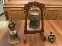 Three brass bells including one with a wooden stand and gong, one smaller bell with a figure handle, and a miniature painted bell.