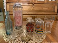 Front view of lot showing a variety of Coca-Cola glasses, blue glass bottle, tall clear plastic straw holder filled with pink straws, and brass items on white lace tablecloth.