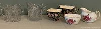Five cream and sugar sets displayed on a flat surface: two small floral china sets and three larger clear cut glass sets lined up