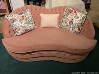 Front view of pink velvet loveseat showing curved back, seat cushion, and three pillows (two floral, one striped).