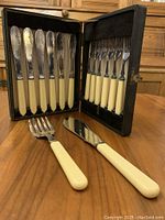 6 fish knives and 6 fish forks in original black case, off-white handles, blades and tines silver plated, plus 2 piece serving set displayed on wooden surface.