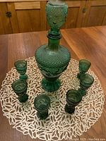 Full set placed on a round white lace doily on a wooden table, showing the decanter with stopper surrounded by six liqueur glasses in emerald green pressed glass.