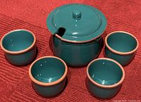 Front angle view of the green glazed soup pot with lid and four matching bowls arranged around it on a red textured surface.