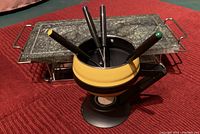 Yellow and brown ceramic fondue pot with four forks inside, on a black burner base, set on a red quilted surface.