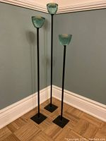 Full view of three handcrafted green glass floor candlesticks on black metal bases against a blue wall and wood floor.