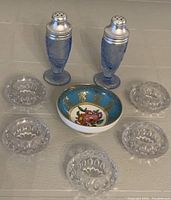 Complete lot showing the two blue glass salt and pepper shakers, five clear crystal salt cellars, and small floral china bowl arranged on a beige fabric surface.