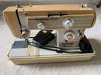 Front angled view of beige and cream electric sewing machine with multiple control dials, foot pedal, and threaded needle.