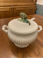 White ceramic soup tureen with ribbed body and lid, one side view showing one handle and part of lid with artichoke handle.