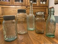 Photo of five vintage glass containers arranged on wooden table in front of wood cabinet. Includes two early Crown sealer jars with black lids, one wide mouth Perfect Seal jar with black lid, a tall embossed decorative bottle with cork, and a green swing top bottle.