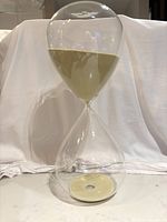 Front view of 15 inch tall glass hourglass sand timer with beige sand in lower chamber, sitting on white surface against white fabric background.