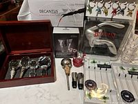 Wide angle shot showing entire barware collection including wooden box with bottle stoppers, glass swizzle sticks, boxed Trudeau screw cap remover, Decantus aerating pourer, plastic shot glasses, and more.