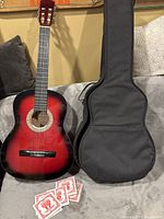 Full view of Denver red acoustic guitar with black soft case next to it, on a couch with gray pillows.