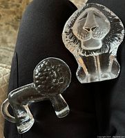 Top view of two clear glass lion sculptures: the smaller Kosta Boda lion lying horizontally and the larger Mats Jonasson lion sitting upright with defined facial features.