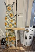 Photo showing ironing board with sunflower print cover, collapsible wood drying rack, and white plastic flexible laundry hamper with handles.
