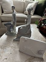 Front view of NAO by Lladro girl figurine seated on rocks holding a jug, new shepherd boy figurine standing and leaning on rock, and a rectangular signed ceramic plaque in front.