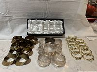 Four sets of napkin rings displayed on a countertop including brass tone diagonal cut rings, shiny gold tone rings, silver tone rings (some wrapped), and a boxed set of clear Rosenthal crystal square napkin rings.