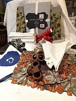 Photo showing new Laura Ashley tablecloth in gray packaging, red plastic and wooden napkin rings, white and blue heart napkins, autumn leaf fabric table runners, and other folded table linens.