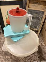 Photo showing the vintage Tupperware pitcher with red lid, two turquoise rectangular containers with lids, and two large white plastic lids beneath them.