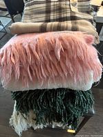 Stack of four blankets showing various textures and fringes: pink mohair, brown plaid wool wrap with visible label, white fleece, and green knit blanket.