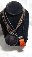 Front view of two necklaces on black display bust, showing large silver disc pendant and coral pendant with leather cords and silver beads
