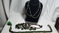 View of 3 green jade necklaces displayed on a black bust and table: one multi-strand jade bead necklace, one Navajo style necklace with large jade discs on brown cords, one long jade bead mala necklace with tassel