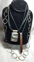 Full view of the three necklaces displayed on a black bust showing a large linked sterling silver ring chain, a geometric pendant on leather, and a wood pendant on silver chain.