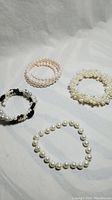 Four bracelets shown on a light fabric background, including white pearl, pink pearl, blackstone with pearls, and single strand pearl bracelet.