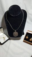 Photo showing all three items together: the silver geometric pendant necklace displayed on a black bust, gold plated initial 'F' pendant in branded box, and gold plated earrings in box.