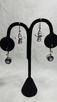 Two pairs of sterling silver earrings displayed on a black velvet stand showing the angel-shaped earrings with blue sapphire and amethyst stones and a pair of purple amethyst rose-shaped studs hanging below.
