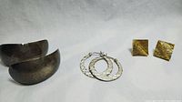 Photo showing three pairs of earrings: large hammered silver half-hoops, medium textured silver hoops, and gold-tone square stud earrings.