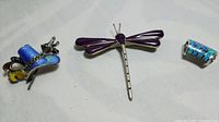 Three sterling silver items including a guilloche enamel hat pin, purple enamel dragonfly pin, and opal paneled pendant shown together on fabric.