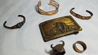 View of three copper bracelets, one with turquoise stone, a western-themed belt buckle, and three rings including a mixed metal bull head ring and two plain rings.