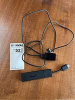 Amazon Fire Stick device with HDMI converter cable and USB charger cable lying on a wooden surface with MaxSold lot measurement card nearby