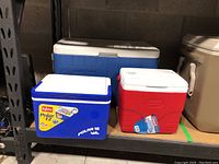 Three coolers on shelf including large blue Coleman, smaller red Coleman, and blue Igloo cooler