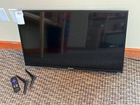 Front view of Hisense 32" TV with remote and two feet placed on carpet.