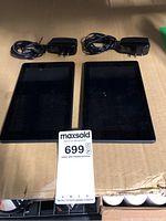 Top front view of two Amazon mini tablets with two charging adapters on a cardboard surface.