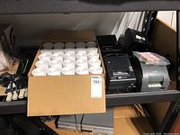 Receipt printers on metal shelving including three black Squirrel printers and one Epson TM-T88V printer with approx. 50 rolls of white thermal receipt paper in cardboard box and power cords.