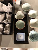Top-down view of six uniquely shaped Chinese porcelain bowls displayed on a black tray with a small blue and white square plate below, alongside cushioned sections of a black fitted storage box