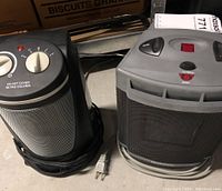 Two space heaters side by side on the floor: a black Holmes heater and a gray AccuTemp by Garrison heater.