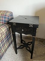 Closed view of ornate black games table with painted floral design on top. Shows locking hinges and drawer front with knob.