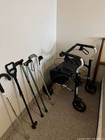 Photo showing tall black and silver Xpresso Lite folding walker alongside five assorted canes and a grabber device lined up against a wall on carpeted floor.