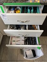 Three open kitchen drawers showing various cutlery and utensils; flatware in top drawer with knives, forks, and spoons; cooking utensils in middle drawer including spatulas, whisks, wooden spoons; bottom drawer with knives and a small white plastic waste basket with black garbage bags.