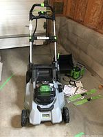 Full assembled mower with handle upright, battery and charger laid out beside it along with grass bag and manuals