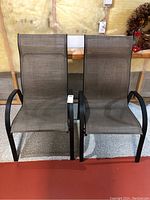 Two matching patio chairs with brown mesh fabric seats and backs, black curved metal frames and armrests, shown side by side on a carpeted floor against a wall.